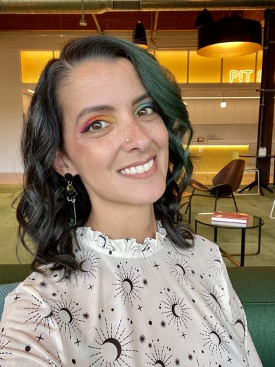 IMG_2604 A photo of Melissa. Melissa has dark hair with green accents. She is wearing UFO earrings, a celestial blouse, and rainbow gradient eyeshadow.